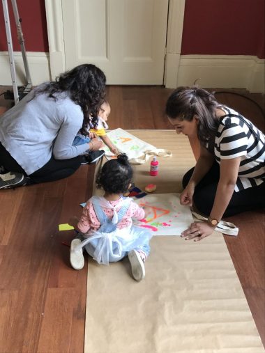 a two women and two young children making art and paint on the ground. The children look no older than 3, and might be a toddler and a baby, there is a jar of fluro paint, and they are working on butcher's paper, making bright geometric shapes and spotches. They appear deep in concentration.