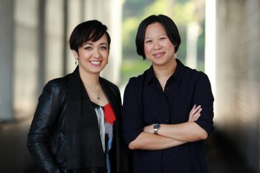 a portrait of Priya Roy and Pearl Town in a semi outdoor setting, they are wearing dark colours, while Priya has a spash of red on her top, they both are smiling at the camera and the photo conveys a grounded and professional presence