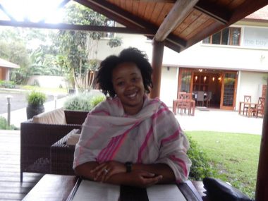 picture of JASMINE MCGINNIS JOHNSON, PHD sitting outside wrapped in shawl, in an outdoor area, there is grass to her left and an open door and a table and chairs that are empty behind her