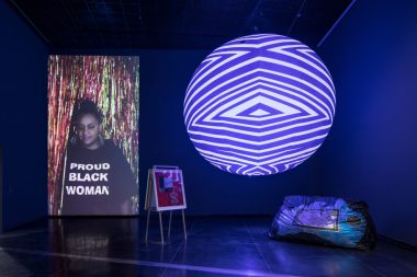 a stage with lights being projected onto a ball, and a picture with a dark skinned woman wearing a tshirt with the words in all caps PROUD BLACK WOMAN, standing in front of a curtain made of shiny, thin, foil streams