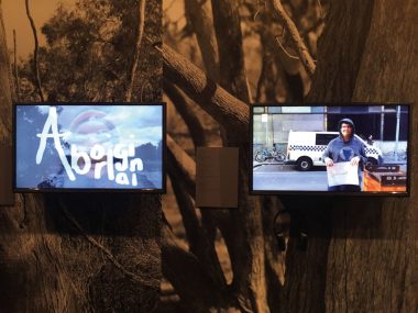 An example of an artwork in the space, has two screens one with the word 'Aboriginal' super imposed over a person wearing a red, yellow, black beanie, while the second screen has a young person in a hoodie in front of a police van holding a sign that simply states 'criminals'