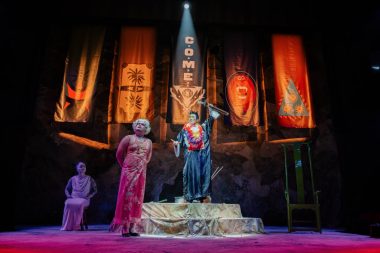 Three performers on stage before a backdrop of coloured banners