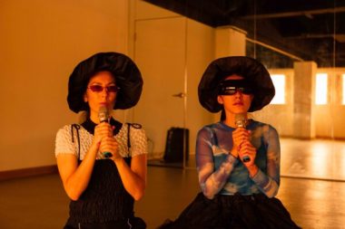 Su Yu Hsin and Angela Goh wearing big black hats and holding mics as part of the work